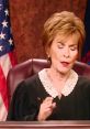 Judge Judy presiding with a serious expression, showcasing her courtroom authority alongside the American flag backdrop.