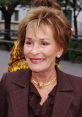 Judge Judy smiles warmly, showcasing her signature style with elegant jewelry and a professional outfit in a vibrant outdoor setting.