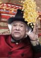 Danny DeVito playfully holds his hand up as popcorn spills around him, dressed in a vibrant red coat and hat, at a fair.