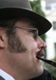 Man wearing a gray hat and glasses, smoking a pipe, showcasing a classic Welsh Chief V1 style with distinctive sideburns.