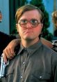 Bubbles from Trailer Park Boys, wearing glasses and a gray shirt, poses seriously with friends in the background.