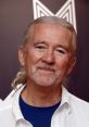 Smiling older man with long hair and a mustache, dressed in a white shirt and blue t-shirt, radiating warmth and charm.