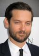 Tobey Maguire smiles confidently in a formal black suit, showcasing his charming demeanor at a public event.