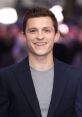 Tom Holland smiles warmly at an event, wearing a navy blazer over a grey shirt, radiating charm and confidence.