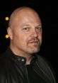 Michael Chiklis with a shaved head, wearing a leather jacket, striking a serious pose against a dark background.