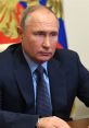 Vladimir Putin seated at a table, looking serious with a focused expression, in front of the Russian flag backdrop.