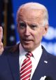 Joe Biden gestures while speaking, wearing a navy suit and red striped tie, with an American flag backdrop.