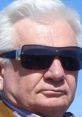 Elderly man with white hair wearing dark sunglasses against a clear blue sky, exuding a confident and relaxed demeanor.