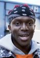 KSI in a casual outfit with a bandana, smiling against an urban backdrop, showcasing his vibrant personality.