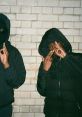 Two individuals in hooded jackets pose against a brick wall, showcasing a street style vibe influenced by UK drill culture.