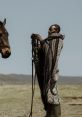 Man in traditional attire prepares horse reins in a scenic landscape, showcasing bond between humans and animals.