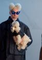 Man with unique hairstyle and blue sunglasses holding a fluffy poodle, styled in a fashion-forward outfit.