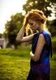 A woman in a blue dress stands in a sunlit park, thoughtful and serene, with flowing red hair and an elegant pose.