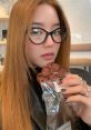 Young woman with long hair holds a chocolate cookie, wearing stylish glasses and dark clothing in a cozy café setting.