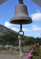 Rustic bell hanging with a rope in a scenic landscape, surrounded by lush greenery and blooming flowers.