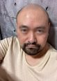 Bald man with a beard in a beige shirt, sitting against a textured wall, showcasing a casual look and style.