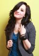 Demi Lovato smiling playfully in a striped blazer and blue top against a yellow backdrop, showcasing her signature style.