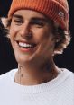 Justin Bieber smiles brightly, wearing an orange beanie and white sweater, showcasing his tattoos and youthful charm.