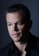 Matt Damon with short gray-streaked hair and a serious expression, wearing a black shirt against a dark background.