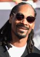 Snoop Dogg smiles confidently, showcasing his signature style with heart-shaped glasses and iconic dreadlocks at a red carpet event.