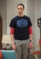 Sheldon Cooper stands in a living room, wearing a blue graphic tee and gray pants, showcasing his quirky personality.