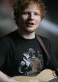 Ed Sheeran performing, wearing a black shirt with a Boba Fett design, playing an acoustic guitar and smiling.