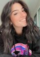 Charli D'Amelio smiling with wavy hair, wearing a colorful hoodie, radiating positivity and charm in a bright setting.