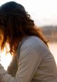 Woman with flowing hair seated by a tranquil waterfront, reflecting on themes of loyalty and connection at sunset.