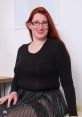 Woman with long red hair and glasses wearing a black top and metallic skirt, posing confidently in a bright room.