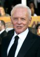 Anthony Hopkins smiles at a red carpet event, dressed in a classic black suit and tie, showcasing his iconic style.