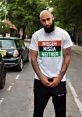 Man wearing a graphic t-shirt with a bold statement, posing confidently on a city street surrounded by greenery.