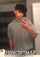 Young Hispanic male taking a selfie in a bathroom mirror, wearing a grey shirt and a chain, captioned "Wednesday.