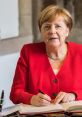 Angela Merkel in a red blazer, writing in a book during a formal event, showcasing her leadership and poise.