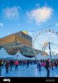 Ice skaters enjoy a winter day at the Birmingham Library, with a Ferris wheel in the background under a clear blue sky.