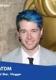 DanTDM smiles at an event, showcasing his signature blue hair and stylish attire, highlighting his vlogger persona.