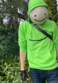 Dream wearing a signature green hoodie with a smiley face, standing among lush greenery, showcasing his iconic style.