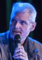 Tom Kane speaking at an event, holding a microphone, with focused expression and distinctive silver hair under blue lighting.