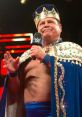 Jerry "The King" Lawler, wearing a crown and royal cape, gestures while speaking on stage at a wrestling event.