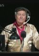 Johnny Gilbert wearing a gold jacket and headset, hosting a game show with a microphone and computer in front of him.