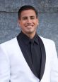 Justin Roberts smiles confidently in a stylish black tie and white tuxedo against a textured gray background.