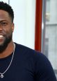 Kevin Hart smiling, wearing a dark shirt and a necklace, in a bright, modern setting with large windows.