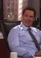 Patrick Warburton smiling in a blue shirt, seated in an office with trophies and a coffee cup in the background.