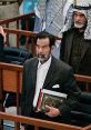 Saddam Hussein stands in court holding a book, surrounded by men in traditional attire, capturing a significant historical moment.