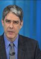 William Bonner delivering news on set, wearing a suit and tie, with a focused expression against a blue background.