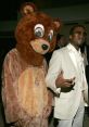 Kanye West in a stylish suit poses with Dropout Bear, showcasing unique fashion and playful spirit at a glamorous event.