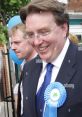 John Michael Howell smiling at an event, wearing a blue rosette, with a cheerful atmosphere and balloons in the background.