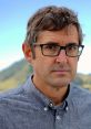 Louis Theroux with a serious expression, wearing glasses and a blue shirt, against a scenic mountainous background.