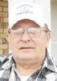 Portrait of an elderly man with glasses, wearing a cap and a plaid shirt, smiling outdoors in a relaxed setting.