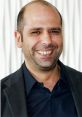 Checco Zalone smiling in a black jacket against a textured white background, showcasing his charismatic personality.