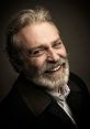 Haluk Bilginer smiles warmly, showcasing his distinct beard and charming personality in a close-up portrait.